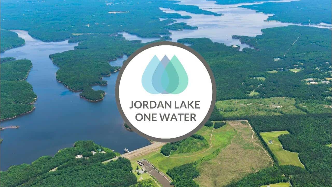 Aerial view of Jordan Lake with the Jordan Lake One Water logo centered over the water and surrounding forest.