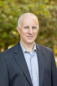Portrait of Matt Day in a suit outdoors with trees in the background.