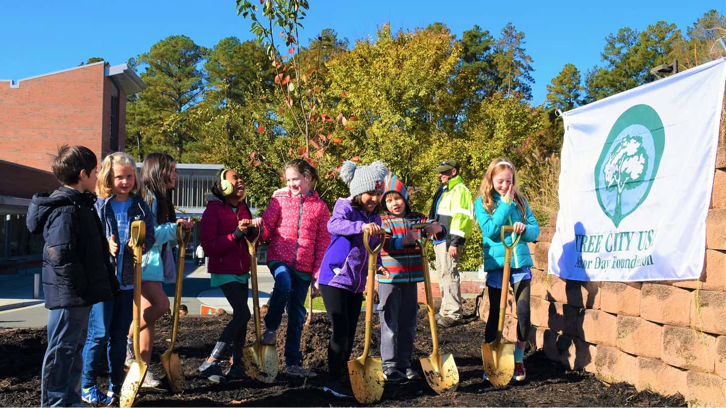 Children holding shovels stand beside a newly planted tree and a Tree City USA Arbor Day Foundation banner.