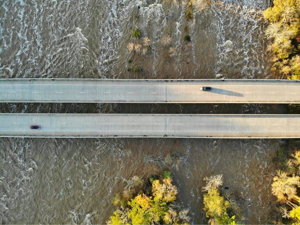 Cars driving on a bridge or a body of water with trees in the background. The image is taken from above.