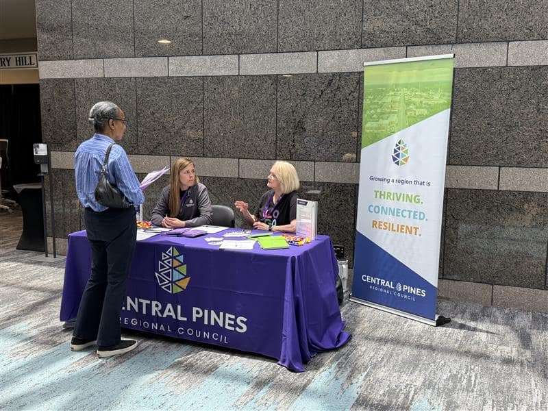 Person standing next to two table where two other people are talking. Central Pines Regional Council logos and media surround the table.