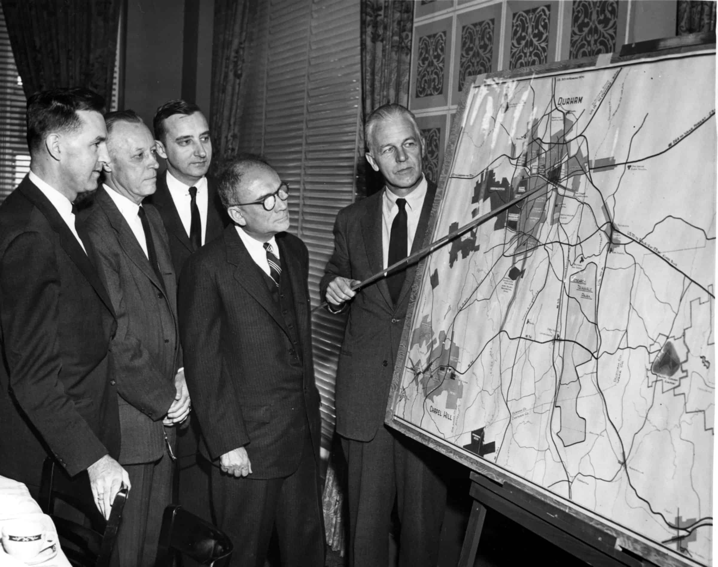 Five members of the Research Triangle Park planning committee examine a regional map showing Durham, Raleigh and Chapel Hill.