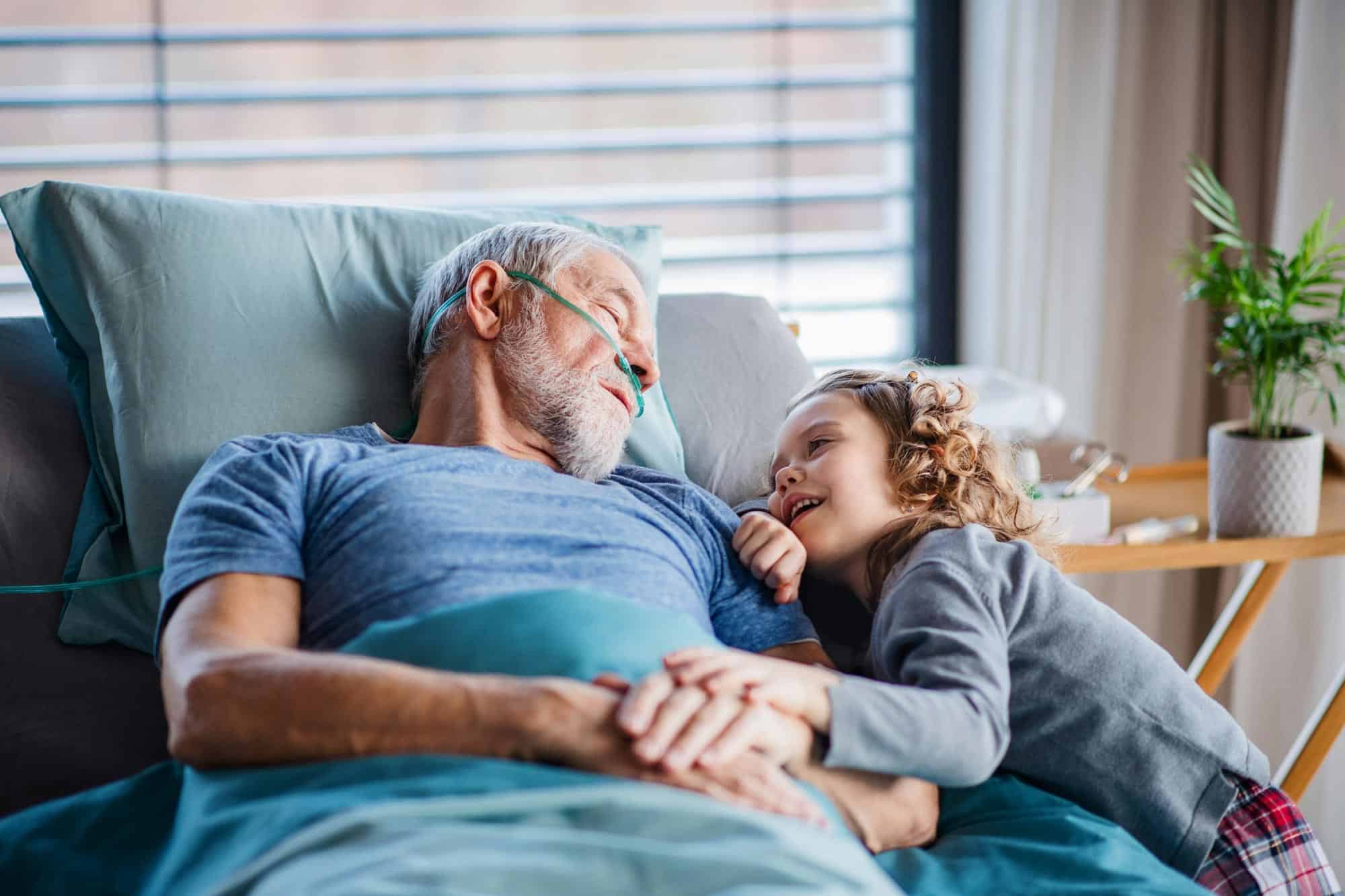 Older person laying head on pillow with young child resting head on the person. They are holding hands.