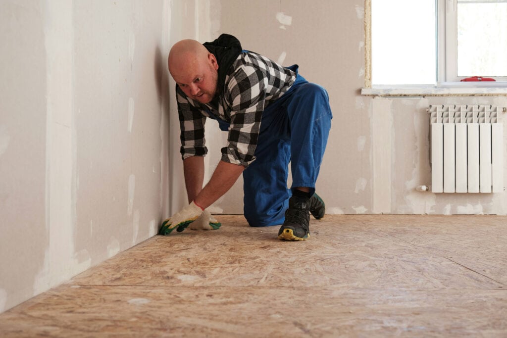 Person kneels on a bare floor next to a wall and uses a tool to plane the floor.