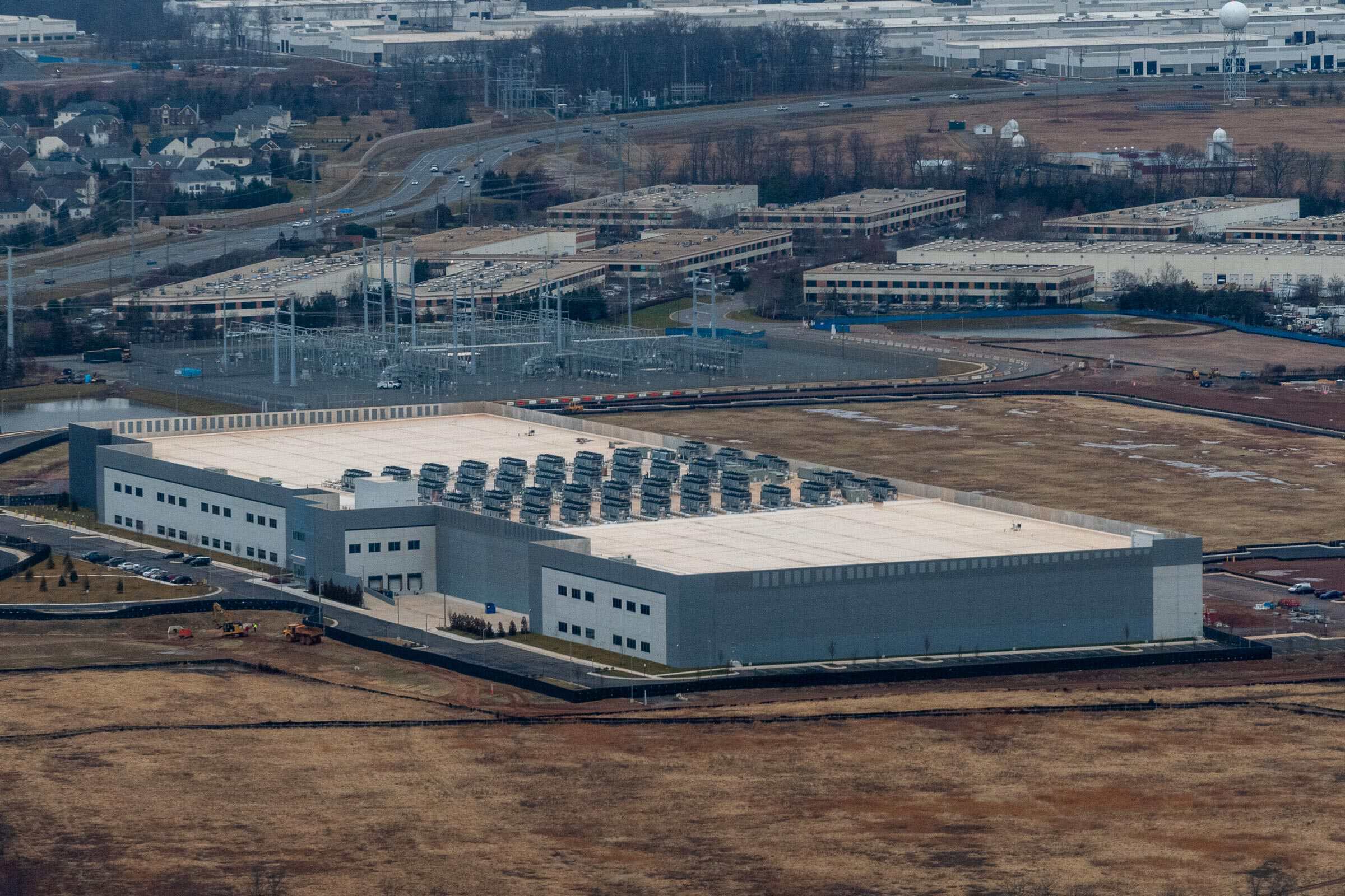 Aerial view of a large data center with rooftop cooling equipment, an electrical substation, nearby industrial buildings and homes.