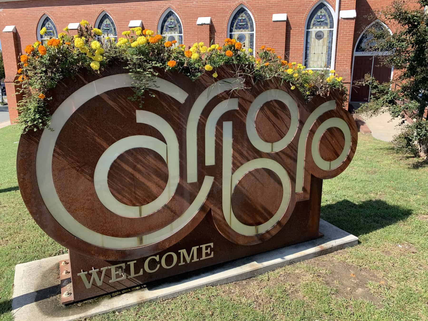 Large metal Carrboro welcome sign topped with colorful flowers in front of a historic red-brick building.