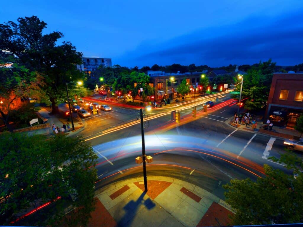 Aerial view of a street intersection from the corner view. Cars zoom past in blurred forms while pedestrians crowd the sidewalks during the evening.