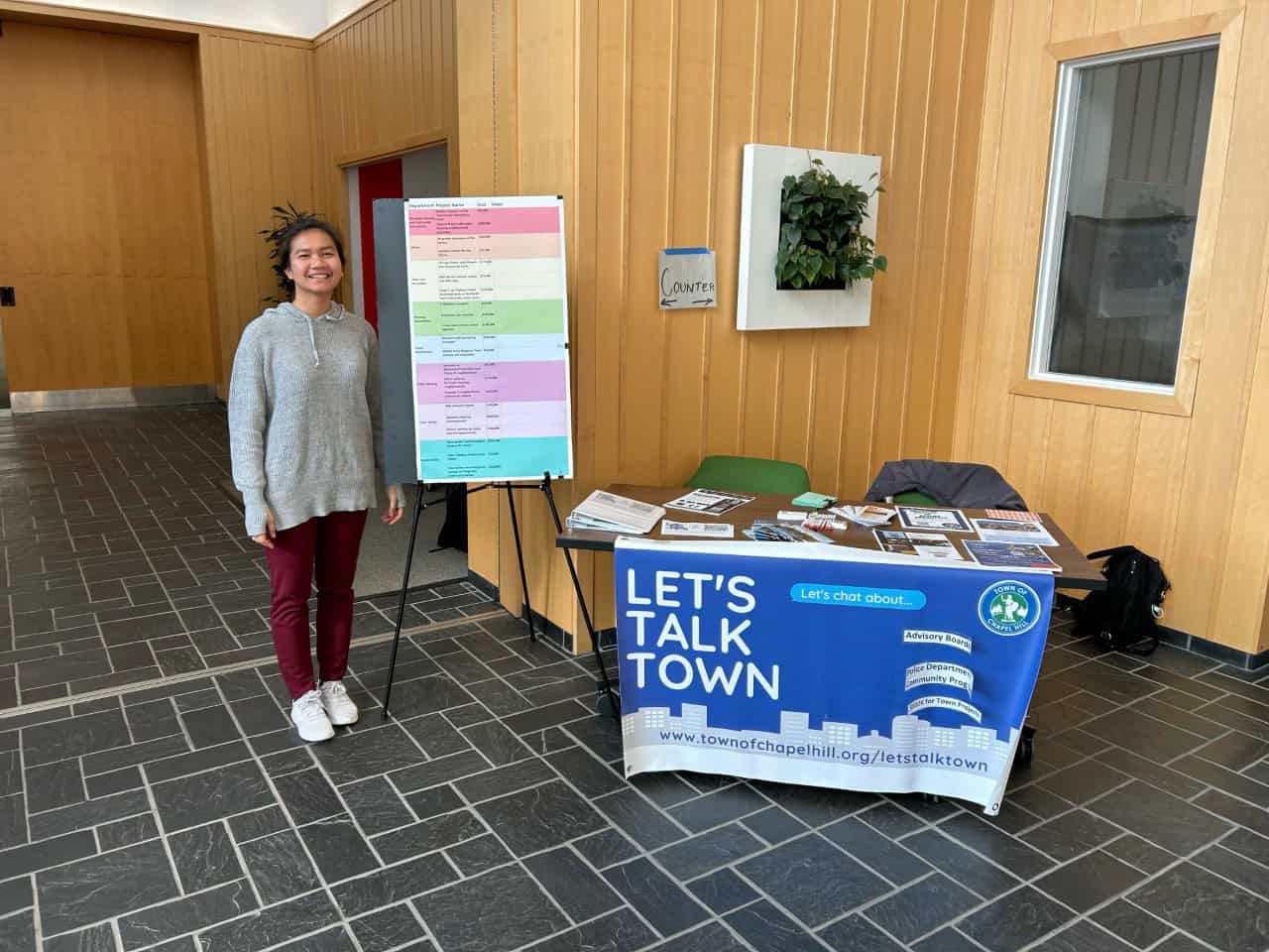 Woman standing beside a Town of Chapel Hill “Let’s Talk Town” outreach table displaying community information and project materials.