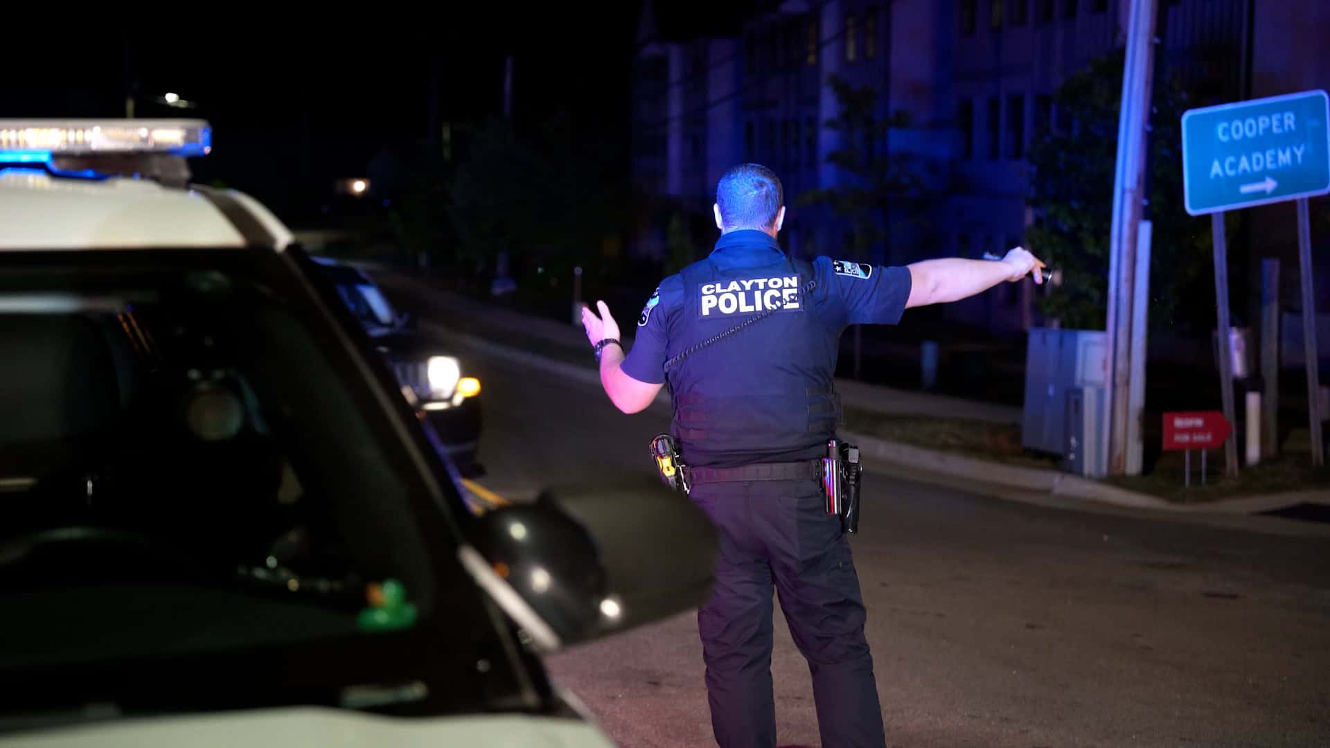 Clayton police officer directing traffic at night beside a patrol vehicle, illuminated by emergency lights near a Cooper Academy sign.