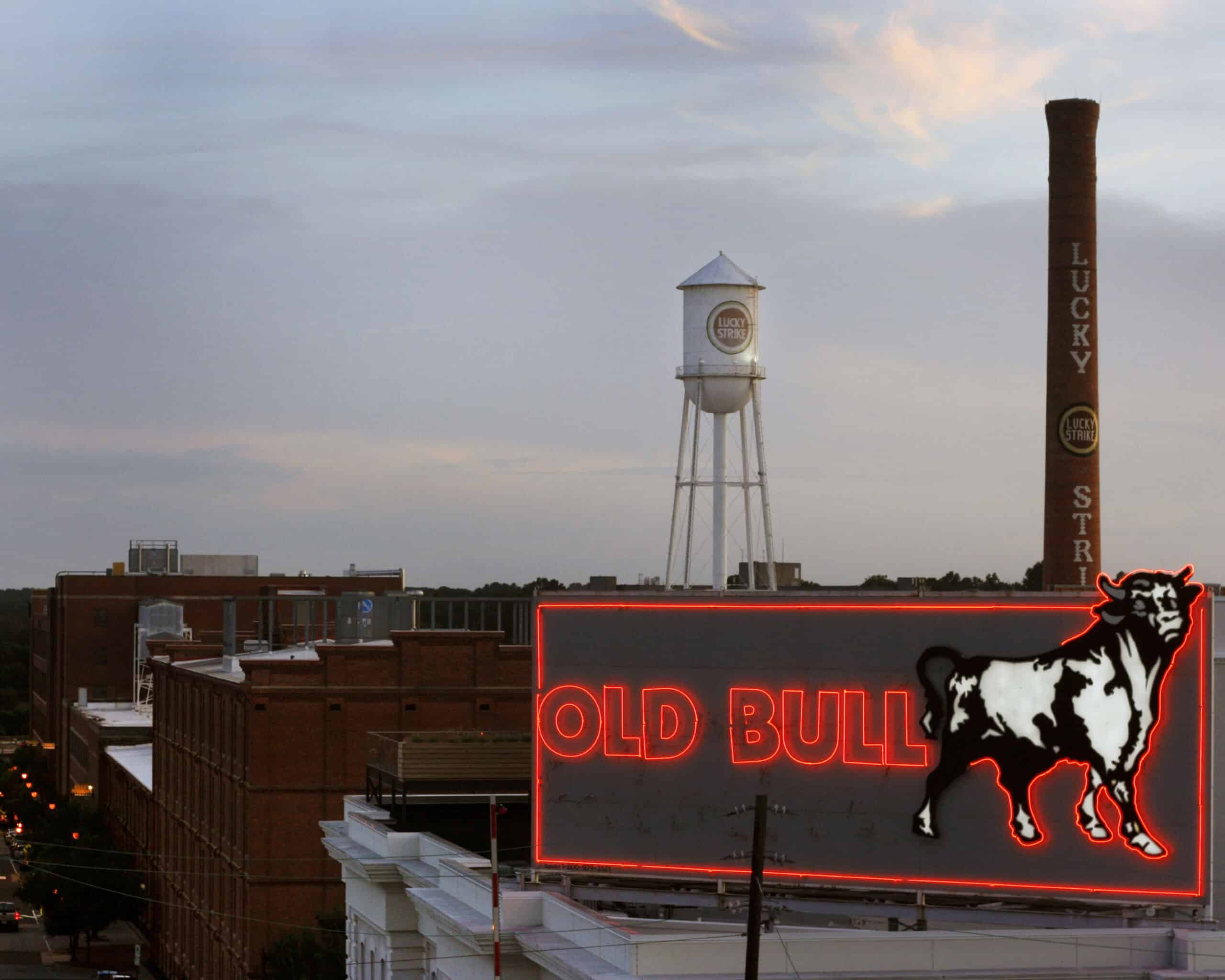 A glowing red Old Bull sign in front of brick buildings, with the Lucky Strike water tower and smokestack behind them.
