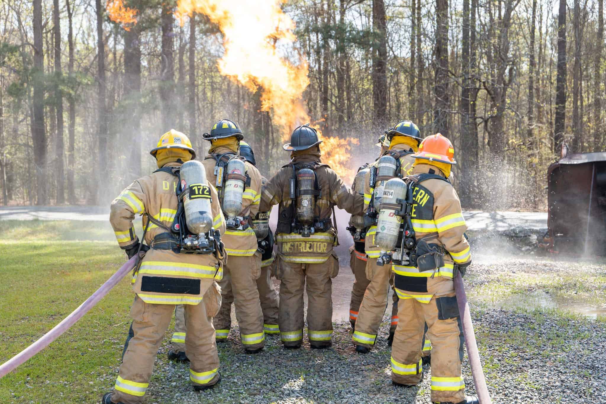 Firefighters in protective gear and breathing equipment stand near flames in a wooded area, holding a hose.