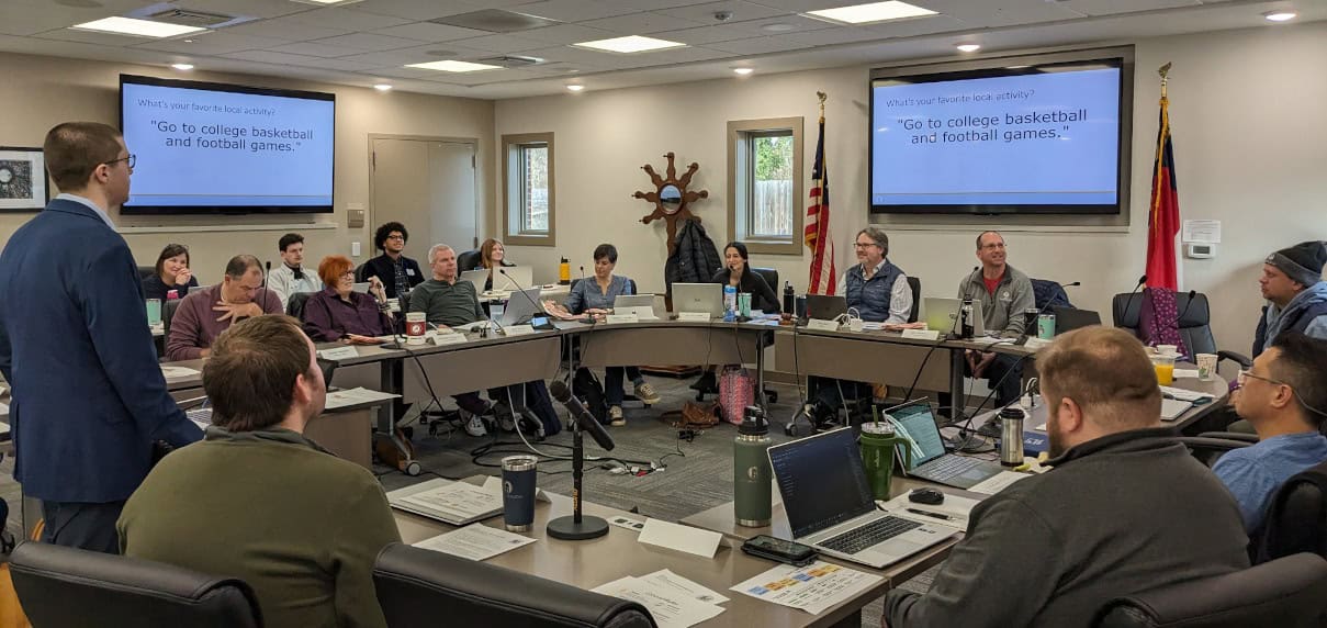 Local officials and attendees seated around conference tables during a Hillsborough Board of Commissioners meeting, with a presenter addressing the room.
