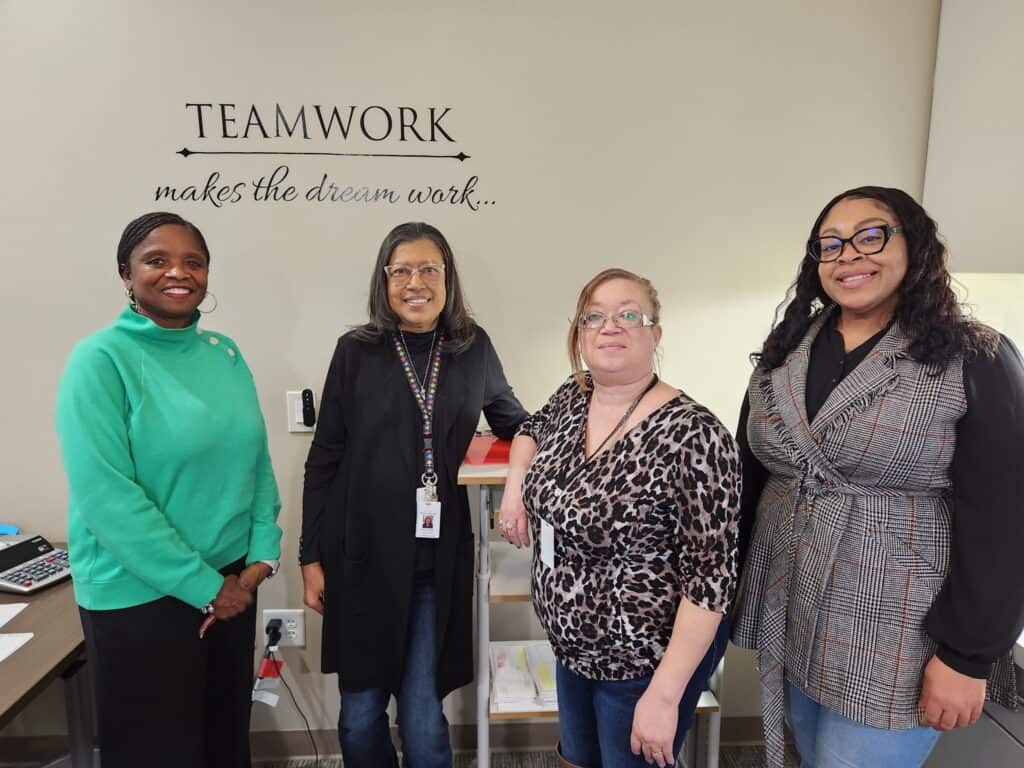 Four Hillsborough employees standing together in an office beneath a wall sign reading “Teamwork makes the dream work.”