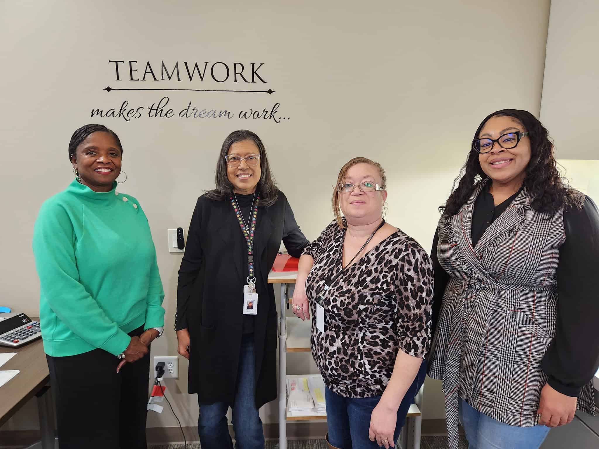 Four Hillsborough employees standing together in an office beneath a wall sign reading “Teamwork makes the dream work.”