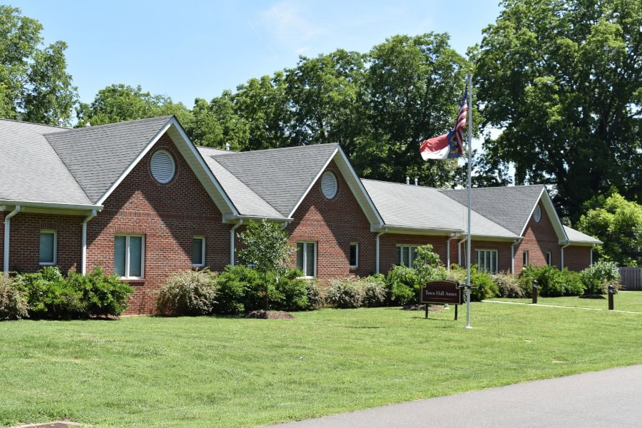Single-story brick Hillsborough Town Hall building surrounded by trees and landscaped grounds, with the North Carolina and U.S. flags flying outside.