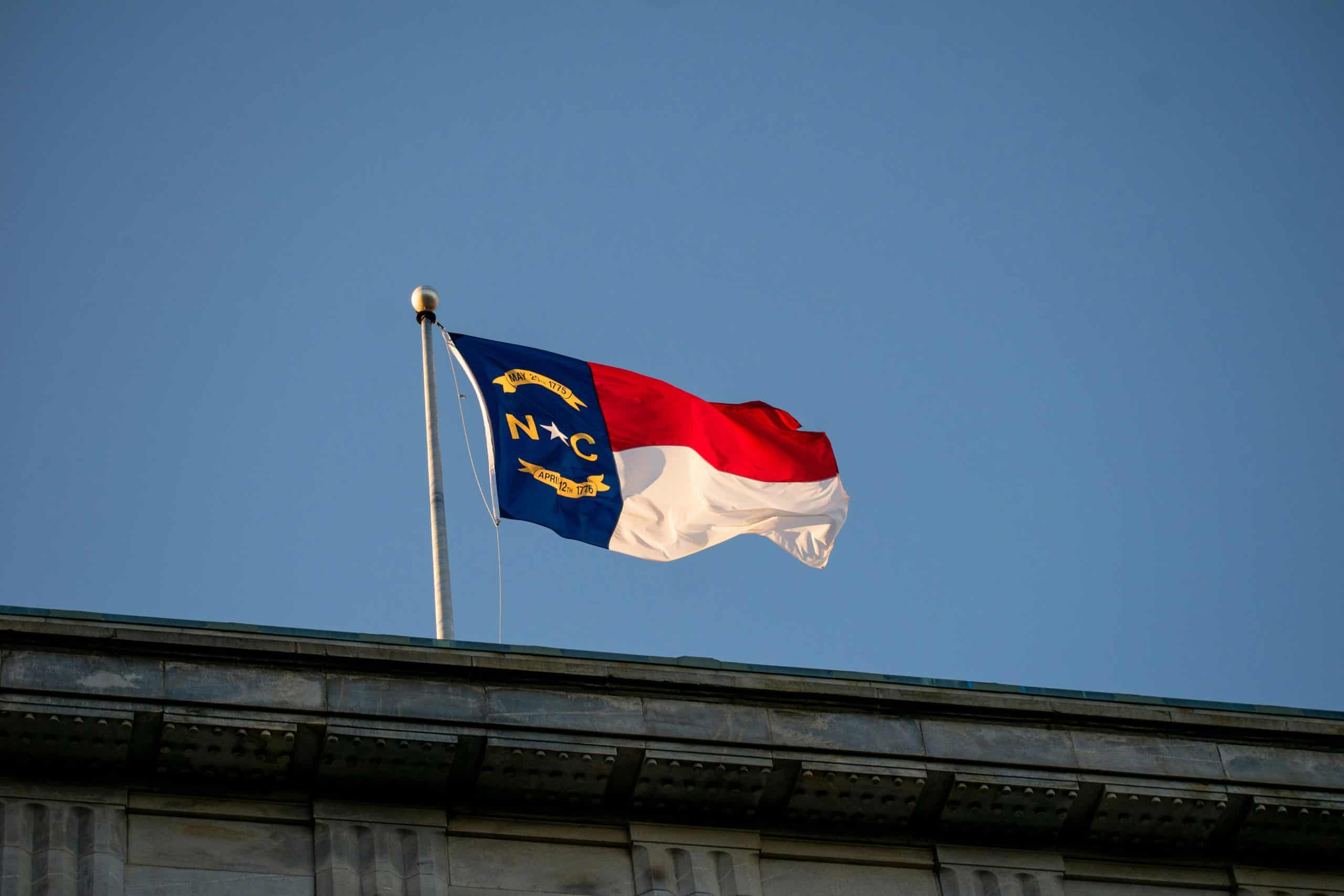North Carolina state flag waving above the roofline of a stone government building against a clear blue sky.