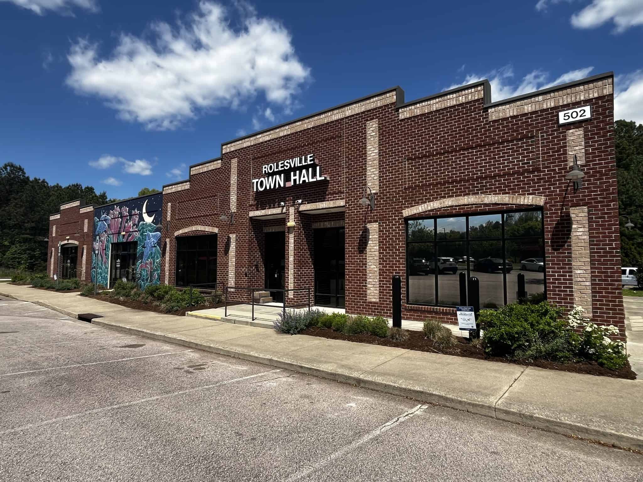 Red-brick Rolesville Town Hall with a colorful mural along one side, landscaped entrance, and blue sky overhead.