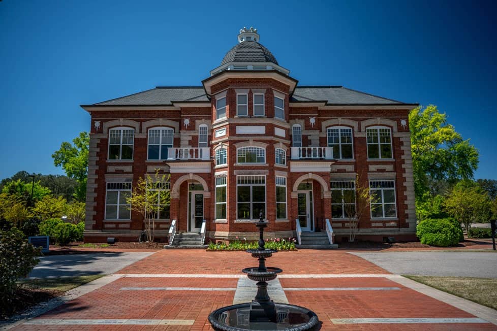 Historic two-story red-brick municipal building with a central turret, arched entrances, and a landscaped brick courtyard with a fountain.