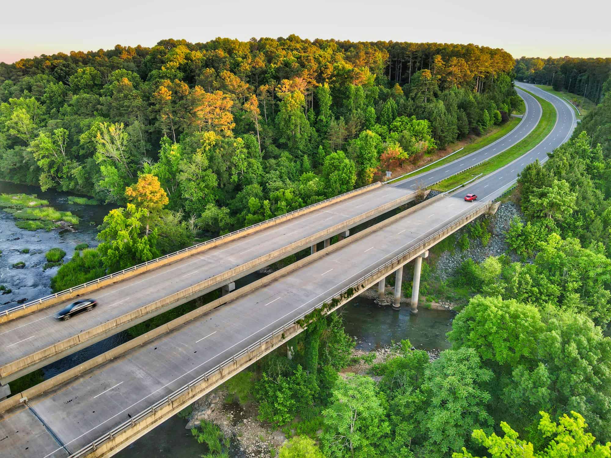 Aerial view of a divided highway crossing a river on two parallel bridges, surrounded by dense green forest in Chatham County, North Carolina.
