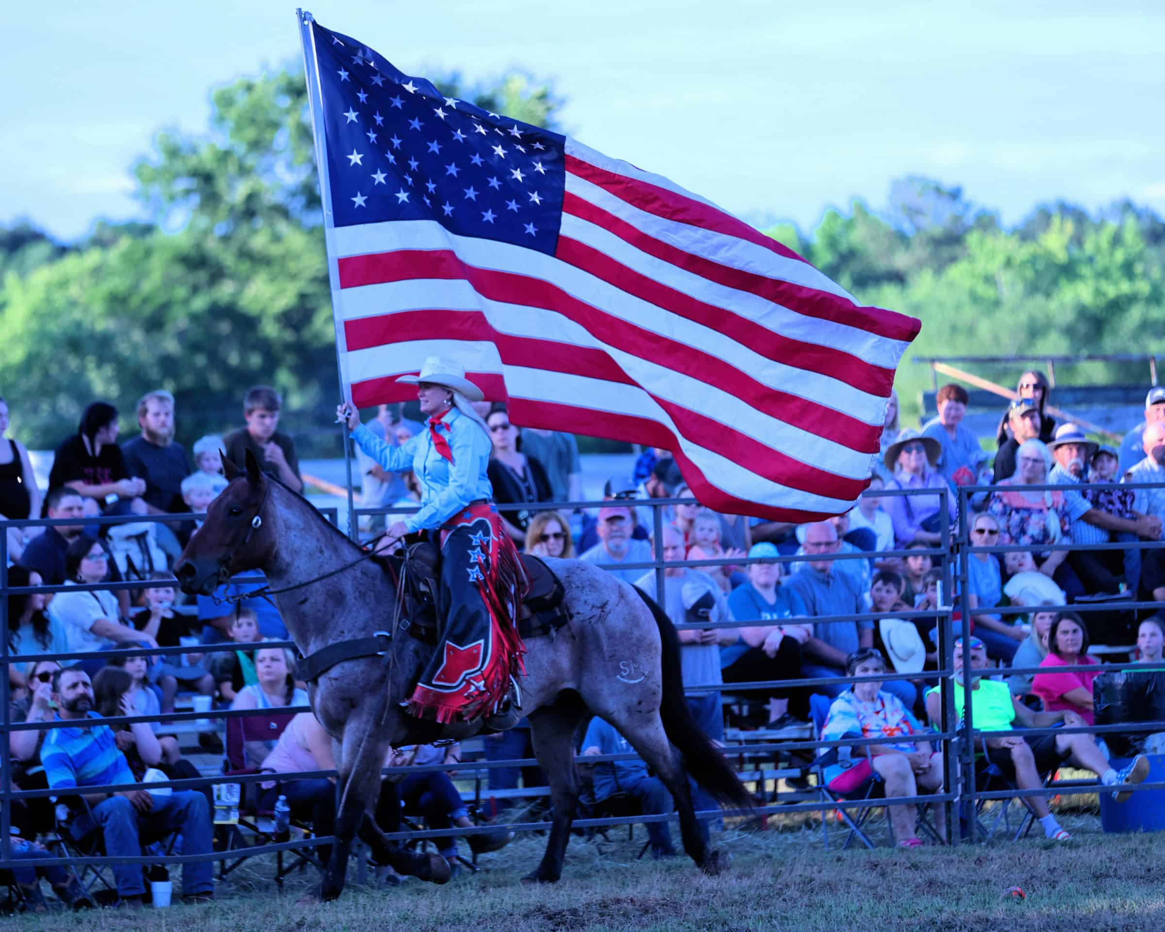 A cowgirl on a gray horse carries a large American flag past spectators seated beside the rodeo arena in Silk Hope