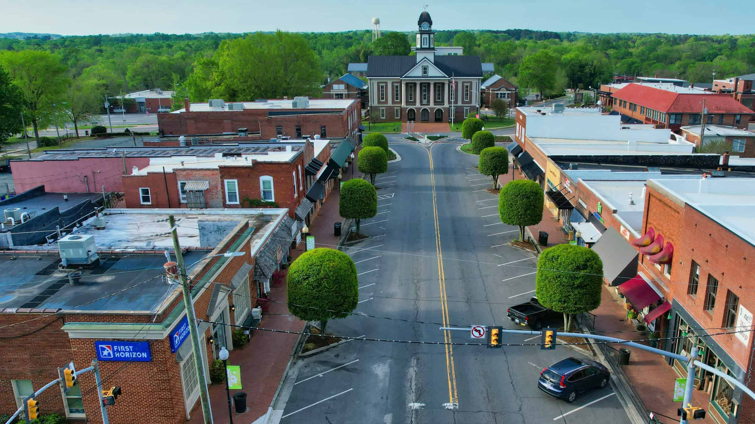 Aerial view of downtown Pittsboro, with brick storefronts and neatly trimmed trees lining the street toward the historic courthouse and clock tower.